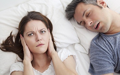 Woman covering ears in bed next to snoring man